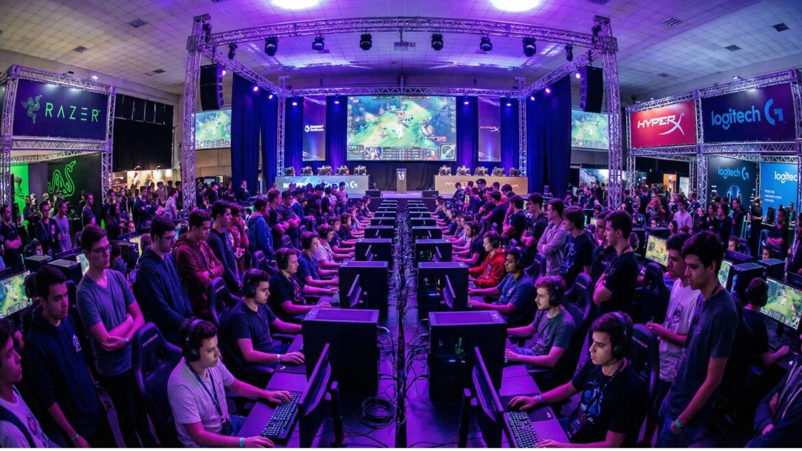 Esports tournament hall at Carolina Games Summit showing rows of gaming setups with competitors and exhibitor booths under dramatic purple and blue lighting
