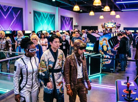 A busy gaming convention hall filled with cosplayers, indie game booths, and retro arcade machines, all illuminated by colorful neon lighting at GeekFest West 2026 in Lynnwood, Washington.