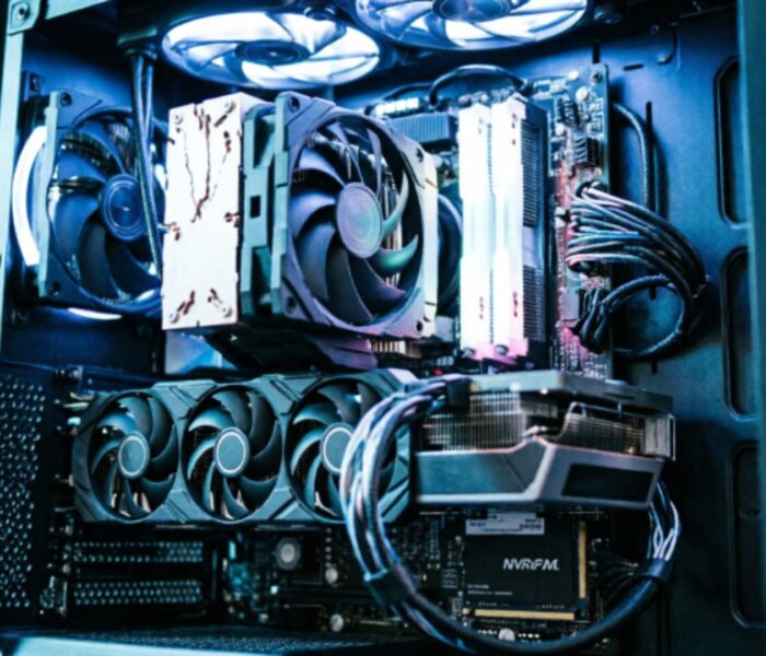 A fully built gaming PC with side panel removed showing GPU, CPU cooler, RAM, and NVMe storage lit with blue and white directional lighting against a black background