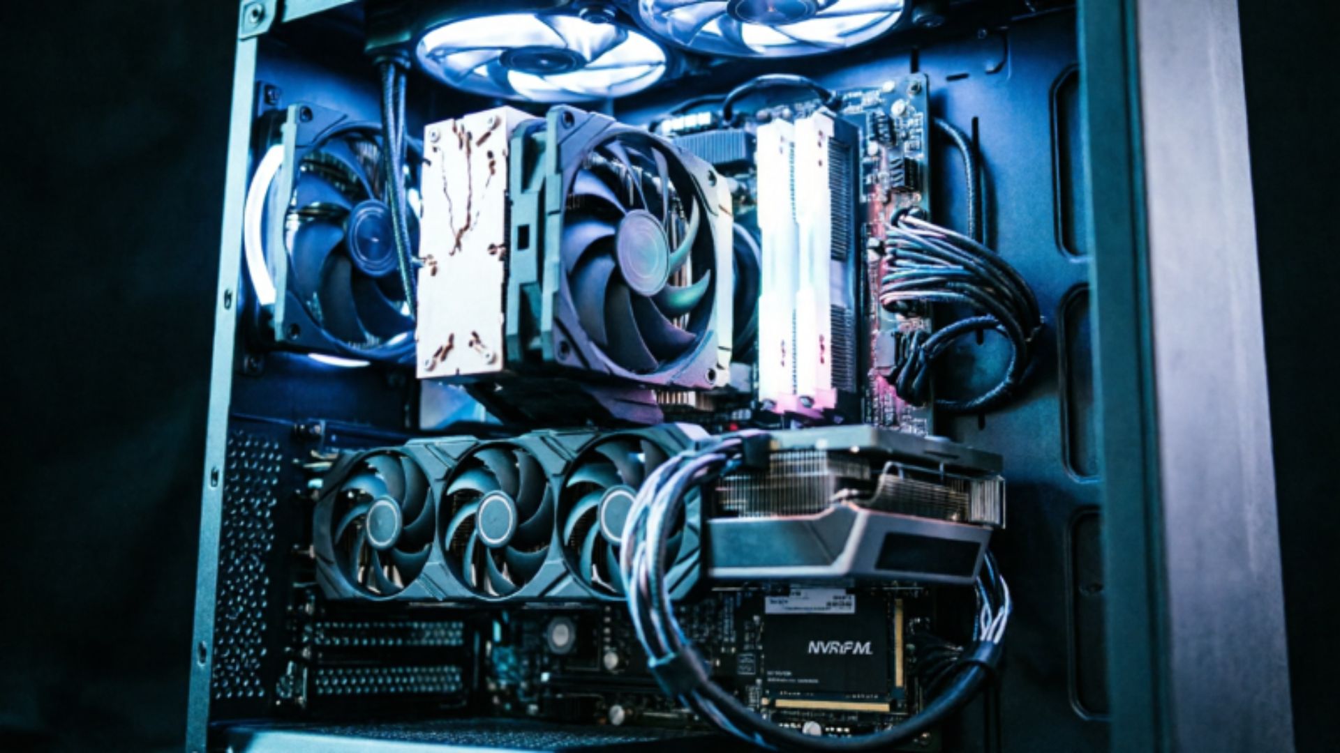 A fully built gaming PC with side panel removed showing GPU, CPU cooler, RAM, and NVMe storage lit with blue and white directional lighting against a black background