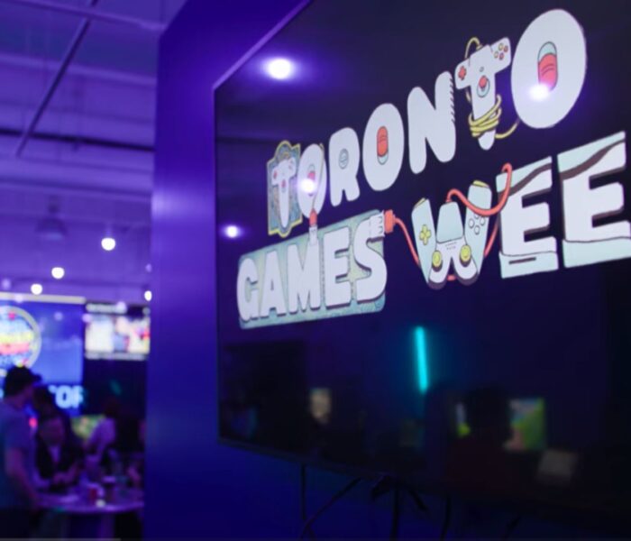 A dimly lit gaming venue with a large “Toronto Games Week” sign, neon lighting, and people gathered around demo stations and tables.