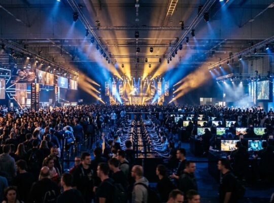 Crowded Gamescom convention hall in Cologne filled with thousands of attendees and illuminated gaming booths under dramatic stage lighting