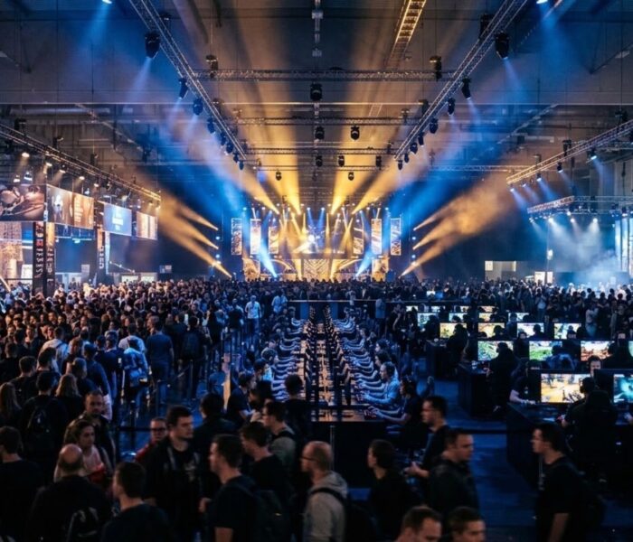 Crowded Gamescom convention hall in Cologne filled with thousands of attendees and illuminated gaming booths under dramatic stage lighting