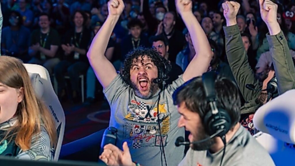 Esports players wearing headsets celebrate an apparent victory at Gamescom 2026, with one player raising both arms as teammates react and a cheering audience watches behind them.